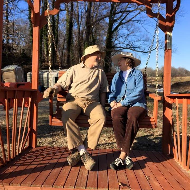 Couple enjoying a sunny day on a wooden swing, sharing a moment of connection and laughter in a serene outdoor setting.