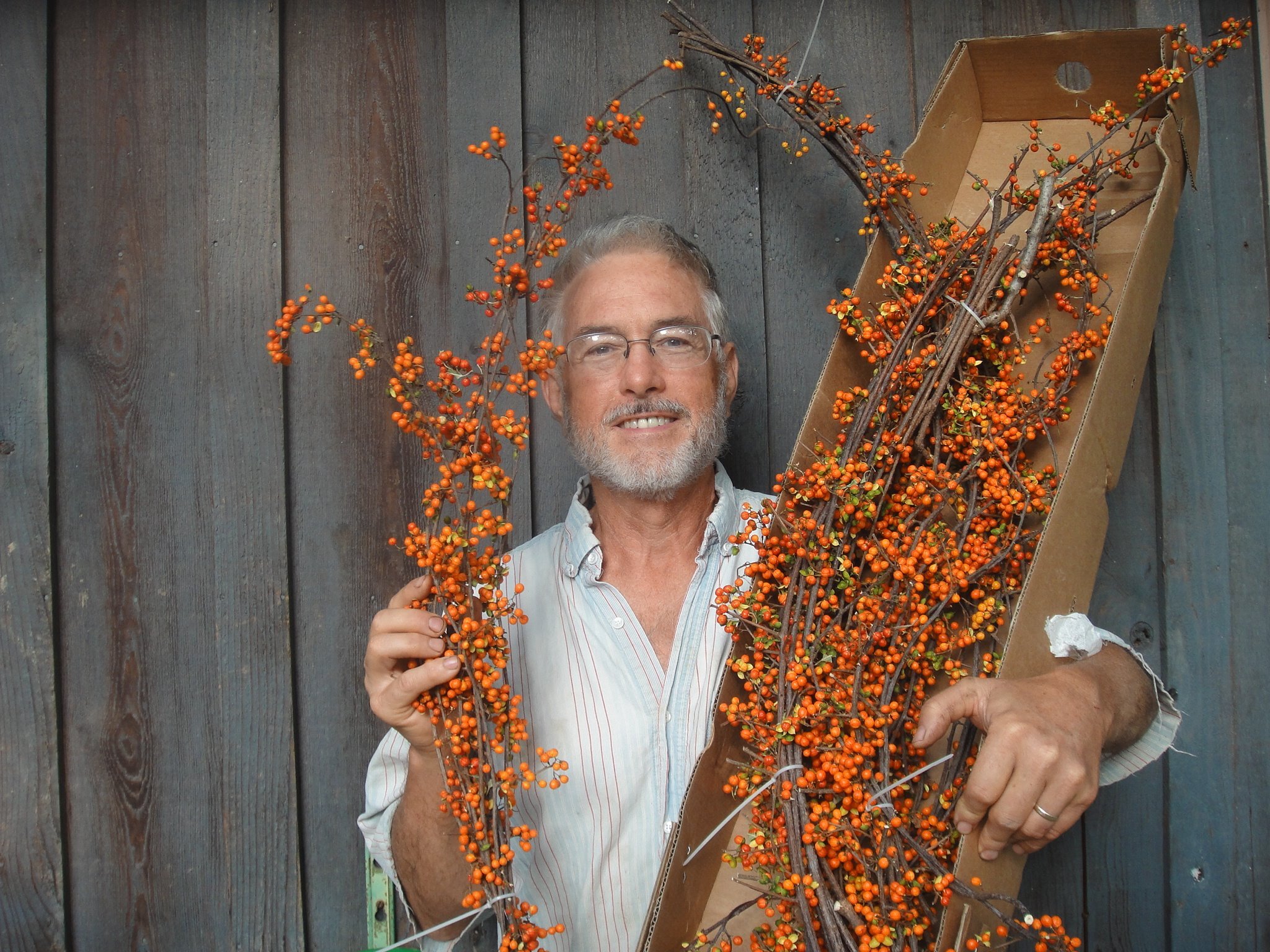 Man holding vibrant orange berry branches in front of a rustic wooden wall, showcasing seasonal floral arrangements.