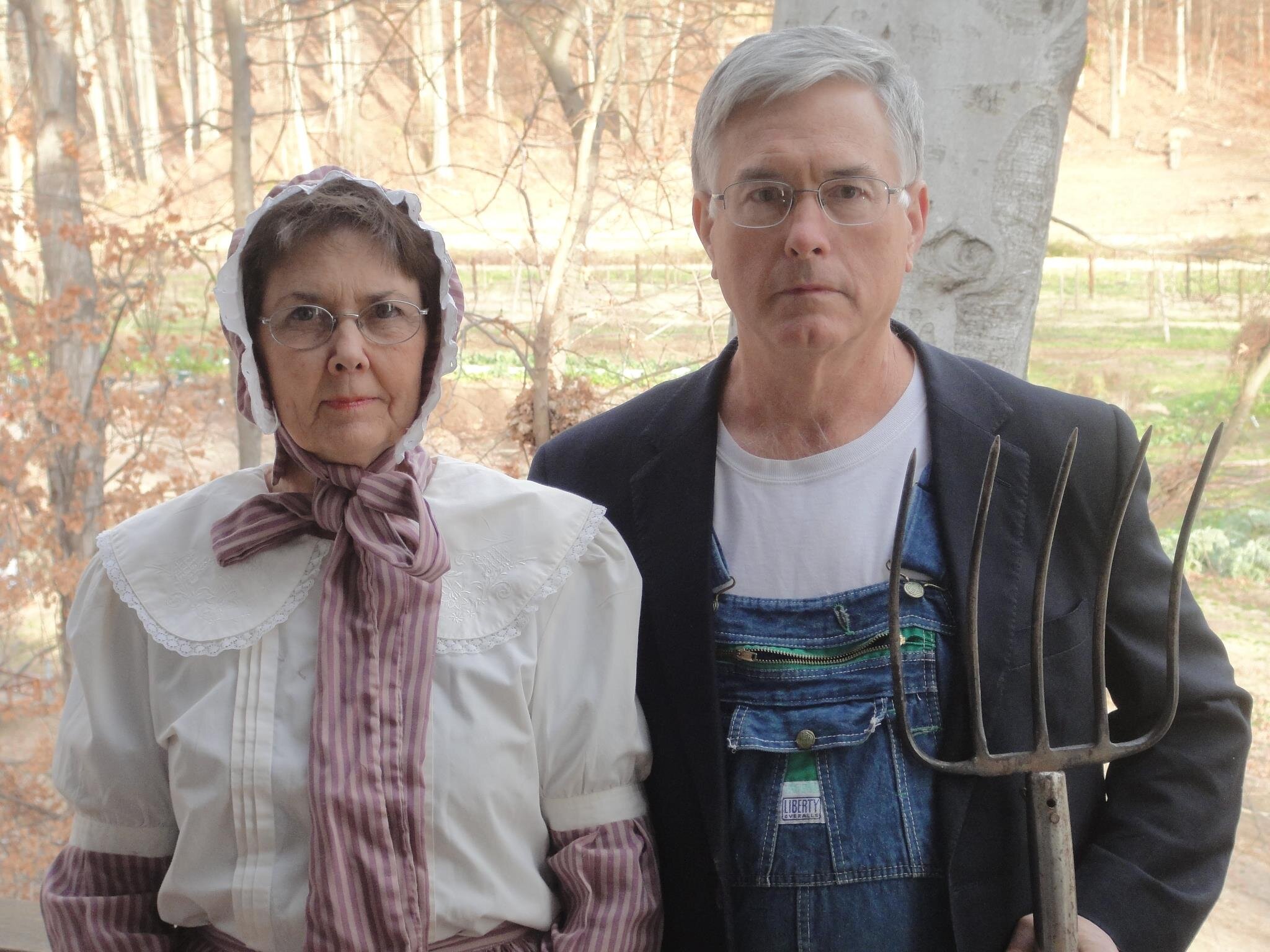 A man and woman dressed in vintage farming attire pose outdoors, with the woman wearing a bonnet and a white blouse with a pink striped ribbon. The man holds a pitchfork and wears overalls with a blazer over a plain shirt. The background features trees and farmland, suggesting a rural setting.
