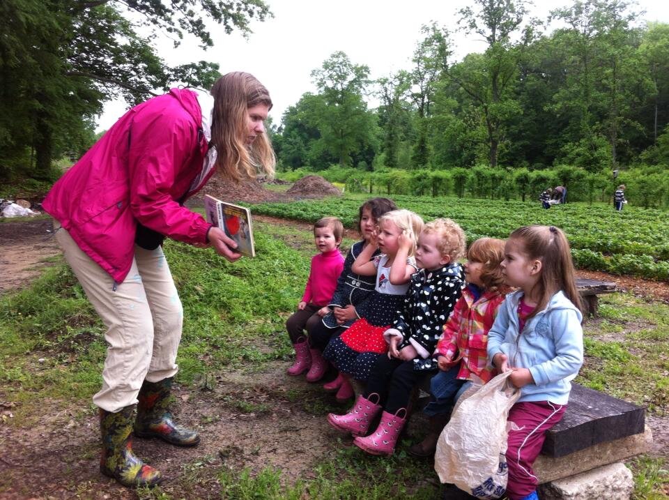Children listening to a lesson outdoors in a garden, with a teacher holding a book. The group is seated on a wooden bench, surrounded by greenery and fields, while other people are working in the background. The children are dressed in colorful rain boots and clothing, engaged and attentive to the instructor.