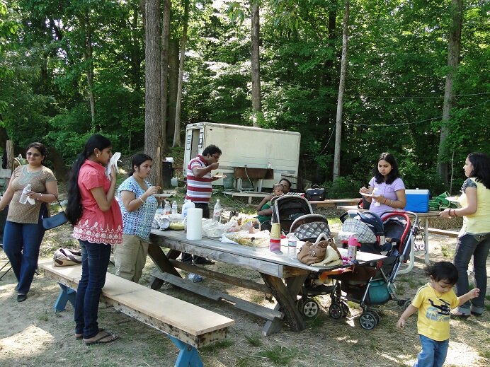 Group of people enjoying a picnic in a wooded area, with a picnic table laden with food and drinks. Children are playing nearby, and some adults are seated or standing, engaged in conversation. Strollers are present, indicating a family-friendly gathering.