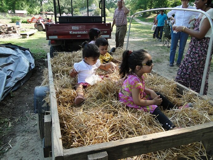Children sitting in a hay-filled trailer at an outdoor event, enjoying a sunny day while adults socialize in the background.