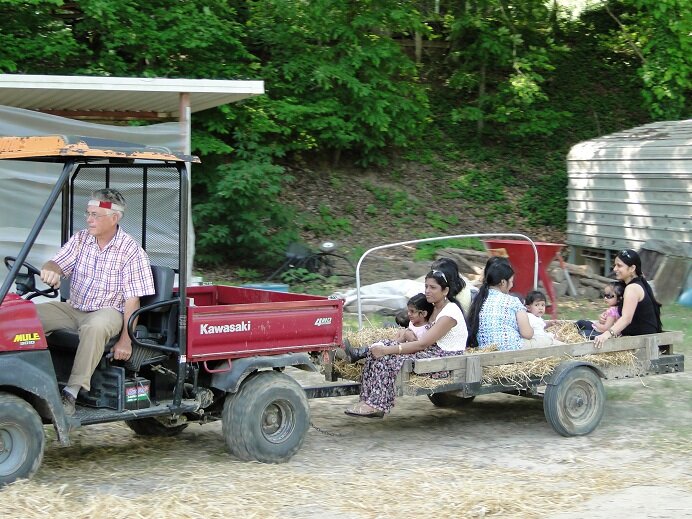 A man driving a Kawasaki utility vehicle pulls a wooden trailer filled with women and children in a rural setting, surrounded by greenery and farm structures.