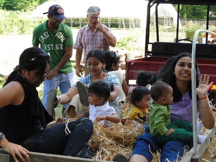 A group of adults and children enjoying a fun day on a farm, sitting in a wagon filled with straw. The scene captures laughter and interaction, showcasing family bonding and outdoor activities.