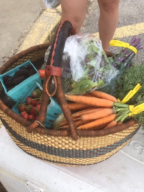 A woven basket filled with fresh organic produce, including carrots, strawberries, blackberries, and mixed greens, resting on a cooler, with a person’s leg visible in the background.