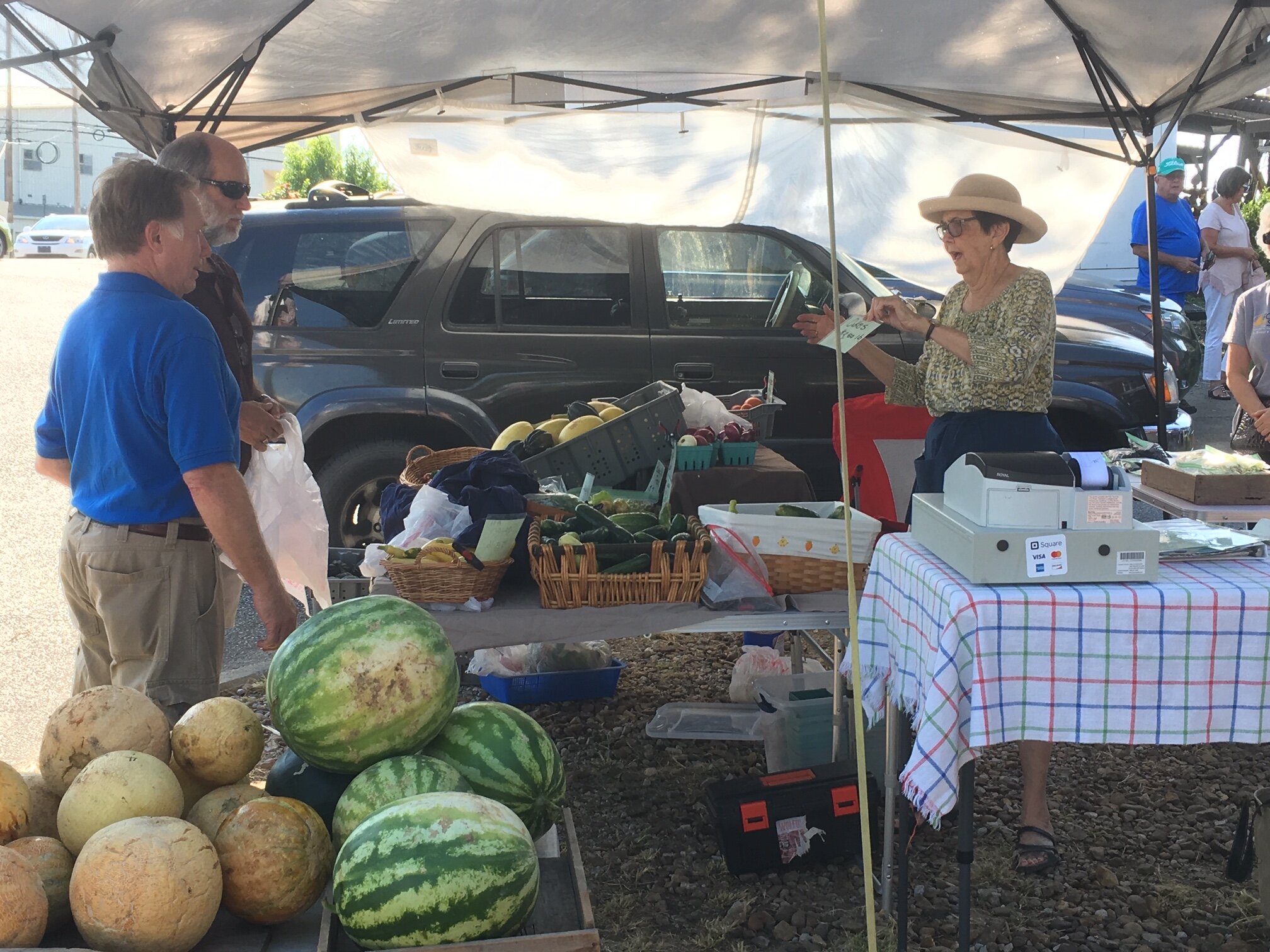 A vibrant farmers market scene showing two customers engaging with a vendor under a tent. The vendor, an elderly woman wearing a hat, is enthusiastically discussing produce, including watermelons and cucumbers, displayed on tables. The background features a parked SUV and other market-goers, highlighting the community atmosphere.