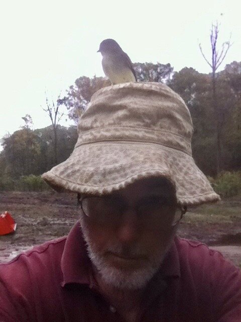 Man wearing a beige bucket hat with a small bird perched on top, set against a natural outdoor background with trees and a cloudy sky.